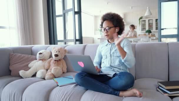 Betrokken bij onderwijs. Vrolijk tienermeisje in koptelefoon met laptop, zwaaiend en lachend tijdens het communiceren met haar leraar tijdens het online studeren. Gelukkig kind zittend op de bank thuis — Stockvideo