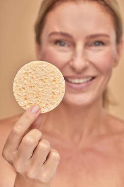 Bej arka planda stüdyoda tek başına dururken elinde makyaj süngeri tutan çekici bir kadının dikey çekimi.