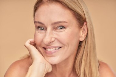 Yaşlanma karşıtı tedaviler. Çekici, mutlu, orta yaşlı bir kadının, yüzüne dokunan ve stüdyoda bej arka planda poz verirken kameraya gülümseyen portresini yakından çek.