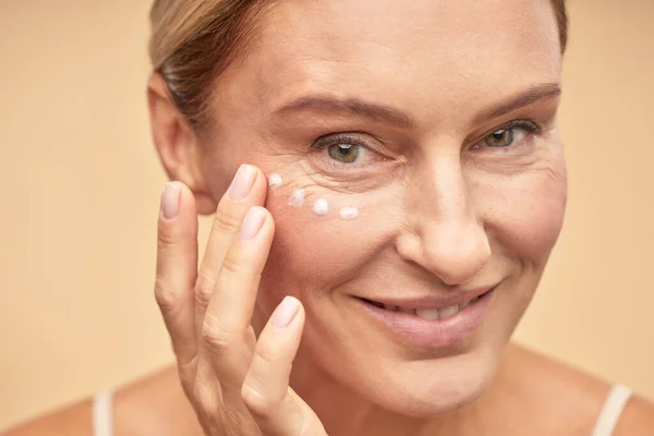 Cuidado de la piel. Primer plano de la hermosa mujer madura aplicando crema antienvejecimiento bajo los ojos, aislado sobre fondo beige — Foto de Stock