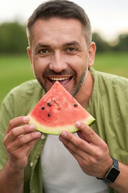 Mutlu genç adam bir yaz günü yeşil parkta karpuz yerken kameraya bakıyor.