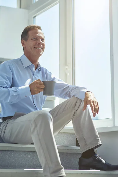 Beristirahat di tempat kerja. Gambar vertikal dari seorang pengusaha dewasa yang bahagia dalam pakaian klasik duduk di tangga di kantor dan minum kopi segar — Stok Foto
