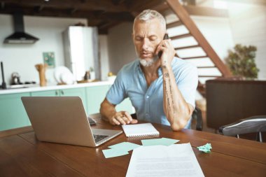 İş konuşmaları. Orta yaşlı, beyaz bir adam masada otururken telefonla konuşuyor ve bilgisayarda çalışıyor.