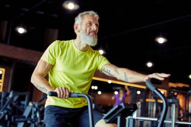 50 'den sonra egzersiz var. Spor kıyafetli olgun bir sporcunun spor salonunda bisiklet sürerken yan görünüşü.