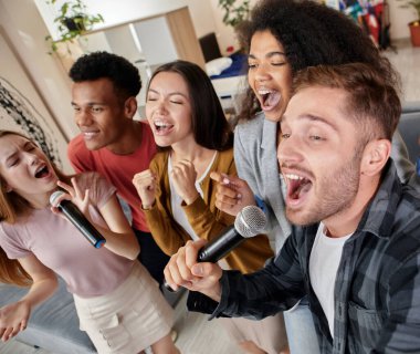 Şarkıyla yüzleş. Evde karaoke yaparken mikrofonla şarkı söyleyen çok kültürlü hevesli arkadaşlar, modern bir dairede duruyorlar.