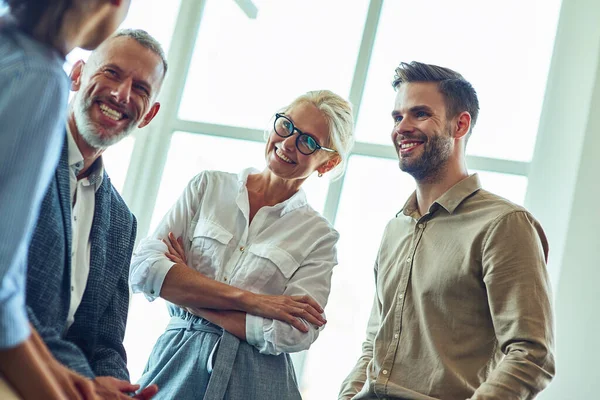 Gente alegre de negocios multirraciales comunicándose, discutiendo algo y sonriendo mientras están de pie en la oficina moderna, trabajando juntos —  Fotos de Stock