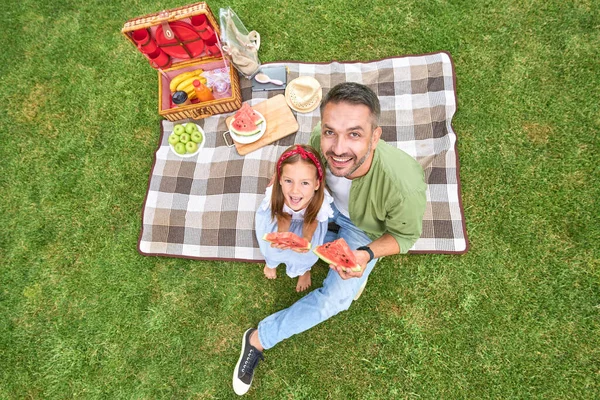Feliz padre y su adorable hijita sonriendo a la cámara, comiendo sandía mientras hacen un picnic en el parque — Foto de Stock
