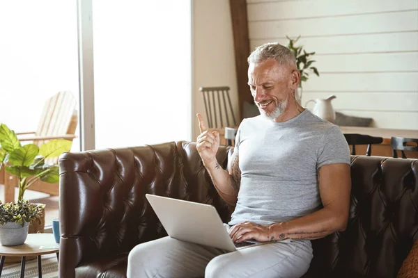 Happy middle aged man in casual closing at laptop and gesturing while sitting on sofa in the living room and working remote — Stok Foto