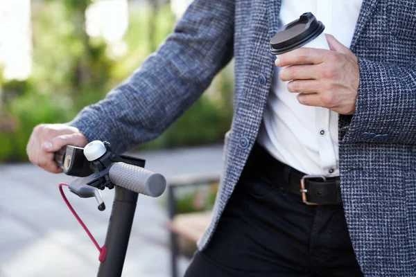 Notwendige Dinge. Großaufnahme der Hände eines eleganten Geschäftsmannes mittleren Alters mit einer Tasse Kaffee, während er tagsüber mit seinem Elektroroller im Freien steht — Stockfoto