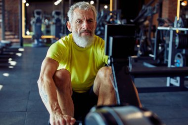 Fitness kulübünde çalışıyorum. Spor giyimli güçlü olgun bir sporcu spor salonunda kürek çekme makinesinde egzersiz yapıyor.