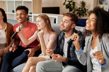 Birlikte şarkı söylemek. Neşeli arkadaşlar evde karaoke yapıyor, oturma odasında kanepede otururken mikrofonla şarkı söylüyorlar.