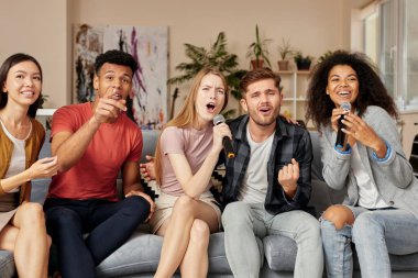 Partine müzik ekle. Evde karaoke yapan, oturma odasındaki kanepede otururken mikrofonla şarkı söyleyen heyecanlı, çok kültürlü genç arkadaşlar grubu.