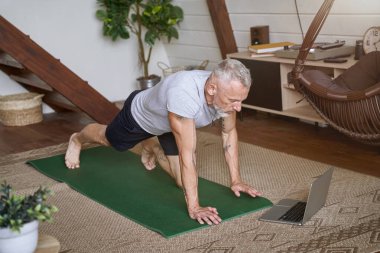 Sportif ve sağlıklı orta yaşlı bir adam modern dairesinin oturma odasında egzersiz yapıyor, dizüstü bilgisayarından online video dersi izliyor.