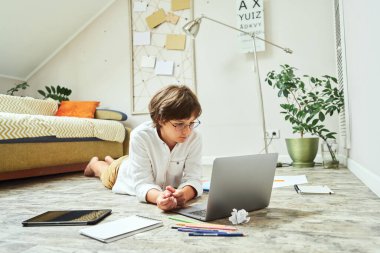 Gözlük takan küçük bir çocuk tahta zeminde yatarken ve dizüstü bilgisayar kullanırken internetten öğreniyor.