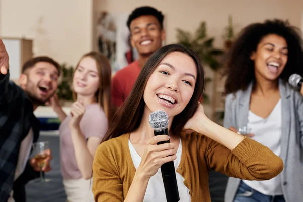 Bawalah sukacita. Wanita Asia muda memegang mikrofon dan bernyanyi sambil bersenang-senang dengan teman-teman di rumah. Sekelompok orang bermain karaoke di rumah. — Stok Foto