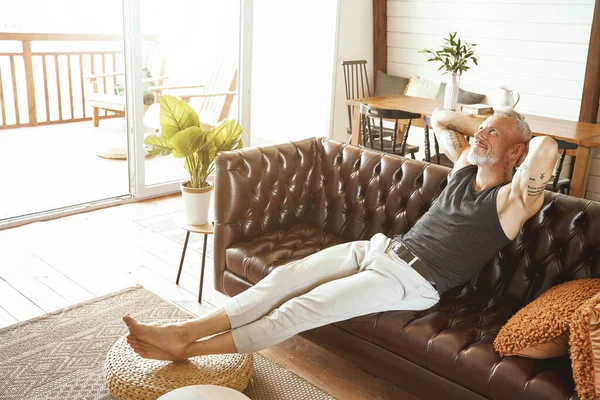 Knappe gelukkige blanke man van middelbare leeftijd in casual kleding ontspannen op een comfortabele bank in zijn moderne appartement, op zoek naar ondersteboven en glimlach — Stockfoto