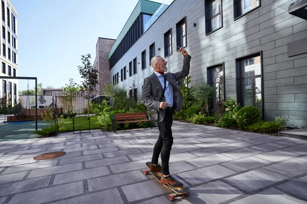 Leben in Bewegung. Ganztägige Aufnahme eines fröhlichen, stylischen, reifen Geschäftsmannes, der lächelt, während er ein Selfie mit seinem Handy macht und mit seinem Skateboard in der Nähe eines modernen Bürogebäudes im Freien fährt — Stockfoto