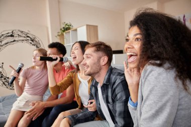 Karaokeye aşığım. Bir grup genç arkadaş eğleniyor, evde karaoke yaparken mikrofonla şarkı söylüyorlar, modern dairede kanepede oturuyorlar.