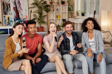 Anlara değer ver. Evde karaoke yaparken mikrofonla şarkı söyleyen, modern dairede kanepede oturan mutlu çok kültürlü arkadaşlar.