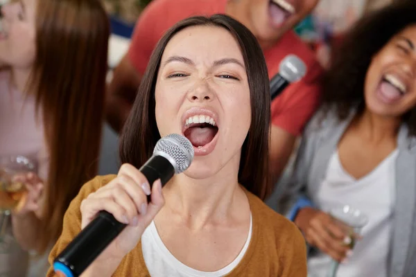 Přines ten zvuk. Detailní záběr krásné mladé asijské dívky drží mikrofon a zpívat při hraní karaoke s nejlepšími přáteli doma. Skupina lidí se spolu baví, pije a jí — Stock fotografie