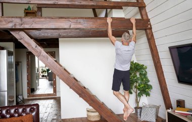 Spor ve sağlıklı orta yaşlı bir adamın evde egzersiz yaptığını, modern dairesinin oturma odasındaki tahta çubukta barfiks çektiğini.