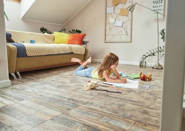 Evde oynayan yaratıcı çocuk. Oturma odasındaki ahşap zeminde uzanmış renkli kalemlerle resim çizen küçük beyaz kız.