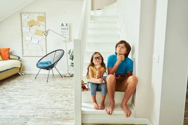 Largura completa de tiro de hermano pequeño y hermana con anteojos sentados en las escaleras en casa y pensando en algo mientras hacen la tarea juntos — Foto de Stock