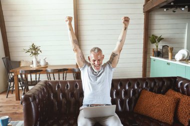 Başarıyı kutluyoruz. Heyecanlı orta yaşlı adam, günlük kıyafetleriyle dizüstü bilgisayara bakıyor ve oturma odasında kanepede oturup uzaktan çalışırken elini kaldırıyor.