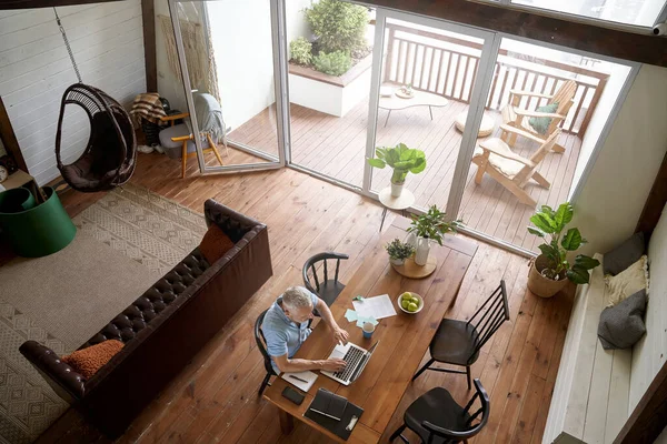 Distance work during self isolation. Top view of a mature caucasian businessman sitting at table in his modern apartment and working online on laptop