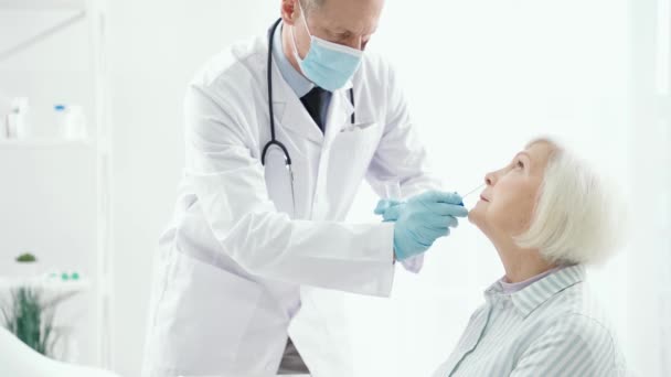 Проверь. Мужчина-врач в медицинской маске берет образец слизи носа у пожилых женщин с помощью медицинского мазка во время проведения процедуры тестирования респираторных вирусов — стоковое видео