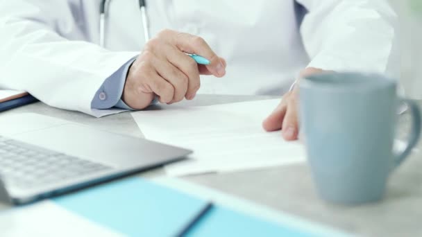 Determining treatment. Close up of hands of middle aged doctor sitting at the desk of his office with laptop, making notes while consulting a patient via the internet — 비디오