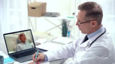 Çevrim içi yardım. Orta yaşlı bir doktor elinde dizüstü bilgisayarla ofisinde oturuyor. İnternet üzerinden üst düzey bir kadın hastayla konuşurken notlar alıyor.