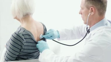 Sağlık kontrolünden. Steteskop takmış, koruyucu eldivenler takmış, kadın hastaları kontrol ediyor, hastanede yaşlı kadınların nefes seslerini dinliyor.