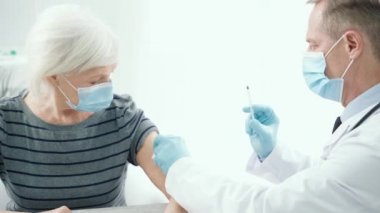 Aşı ol. Tıbbi maske ve eldiven takan erkek doktor hastanede yaşlı bir kadına iğne yapıyor.
