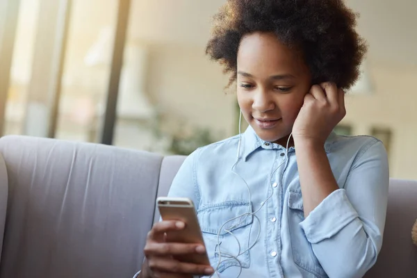 Potret gadis ras campuran memakai earphone, duduk di sofa dan menggunakan ponsel sambil menghabiskan waktu di rumah — Stok Foto