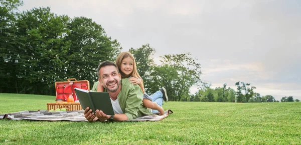 Neşeli küçük kız babasıyla vakit geçiriyor, parkta piknik yaparken kitap okuyor.