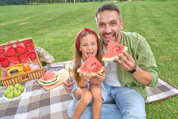 Feliz padre y su linda hijita mirando la cámara mientras come sandía en el parque en un día de verano — Foto de Stock