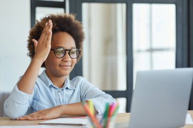 Nişanlı ol. Coşkulu karışık ırk gözlüklü genç kız video dersi sırasında evden ders çalışırken elini kaldırıyor.