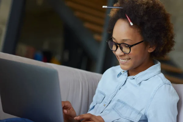 Murid yang sukses. Gadis sekolah remaja yang gembira berkacamata dengan pensil di rambutnya tersenyum sambil menggunakan laptop untuk belajar, duduk di sofa di rumah — Stok Foto