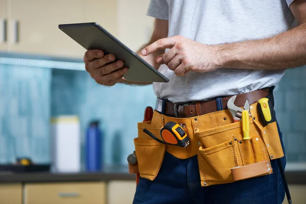 Kapalı alanda dururken dijital tablet kullanan, alet kemeri takan genç bir tamircinin kırpılmış görüntüsü.