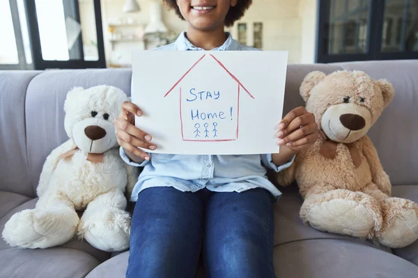 Cropped shot of cute mixed race teen girl in casual clothes smiling at camera and holding Stay Home drawing while sitting on the couch at home during quarantine