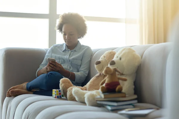 Moderne Kindheit. Fröhlich gemischtes Teenie-Mädchen sitzt auf der Couch und benutzt Mobiltelefon, während sie Zeit zu Hause verbringt — Stockfoto