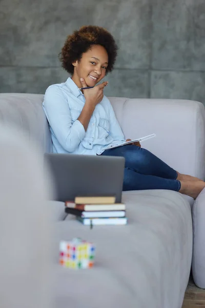 Gadis sekolah remaja ras campuran yang lucu tersenyum di depan kamera sambil mengerjakan PR, duduk dengan laptop dan buku-buku di sofa di rumah — Stok Foto