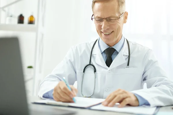 Médico alegre fazendo papelada e usando laptop na clínica — Fotografia de Stock