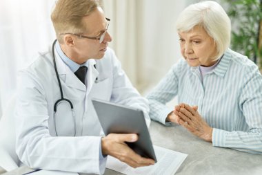 Yaşlı kadınla konuşurken tablet bilgisayar kullanan bir erkek doktor.