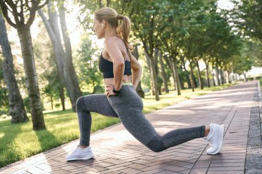 Formda hissediyorum. Spor kıyafetli orta yaşlı bir kadın odaklanmış görünüyor, güneşli bir günde yeşil bir parkta egzersiz yaparken bacaklarını esnetiyor.