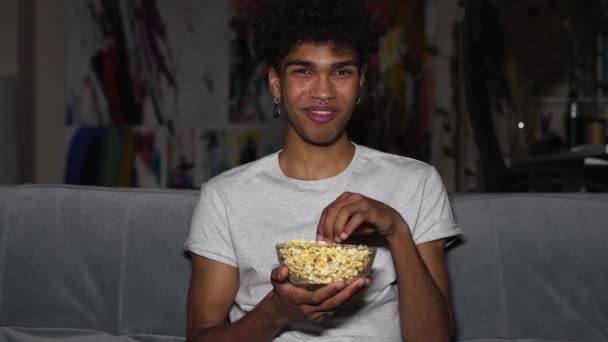 Vermaak. Jonge aantrekkelijke man lachen tijdens het kijken naar comedy film en eten, vasthouden popcorn kom, zitten op de bank in de woonkamer thuis alleen 's avonds laat — Stockvideo