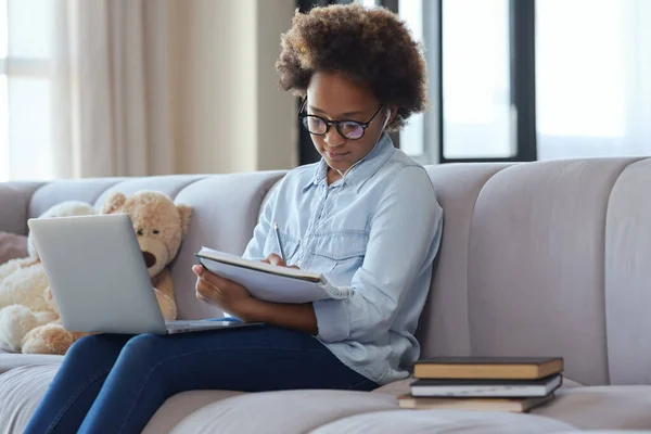 School at home. Teen schoolgirl wearing earphones making notes in her notebook while studying using laptop, sitting on the couch at home