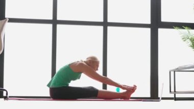 Fitness. Çevrimiçi koç dinlerken neşeli görünen sağlıklı olgun bir kadının yan görüntüsü oturma odasındaki yoga minderinin üzerinde vücudunu esnetiyor.