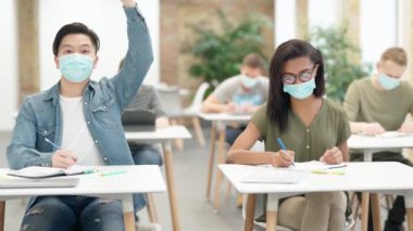 Hevesli bir öğrenci. Üniversitede masada oturan, koruyucu yüz maskesi takan, dinleyen ve notlar alan çeşitli öğrenciler. Sınıfta ders sırasında elini kaldırdı.